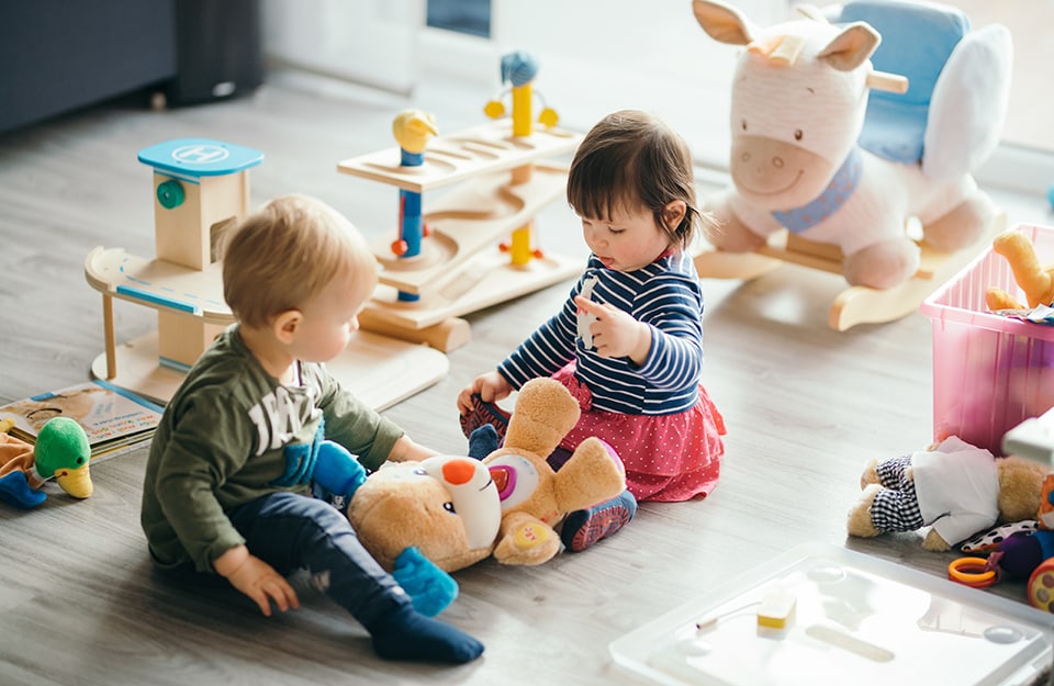 Un bambino e una bambina stanno giocando con un grande peluche sul pavimento in parquet di un asilo nido, circondati da altri giochi e costruzioni in legno