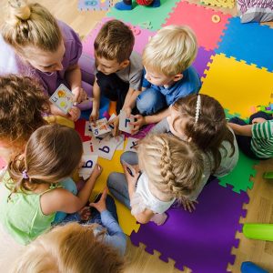 Vista dall'alto di un gruppo di bambine e bambini dell'asilo nido raggruppati in cerchio con una maestra sul parquet della classe, accanto a dei tappetini colorati a forma di puzzle