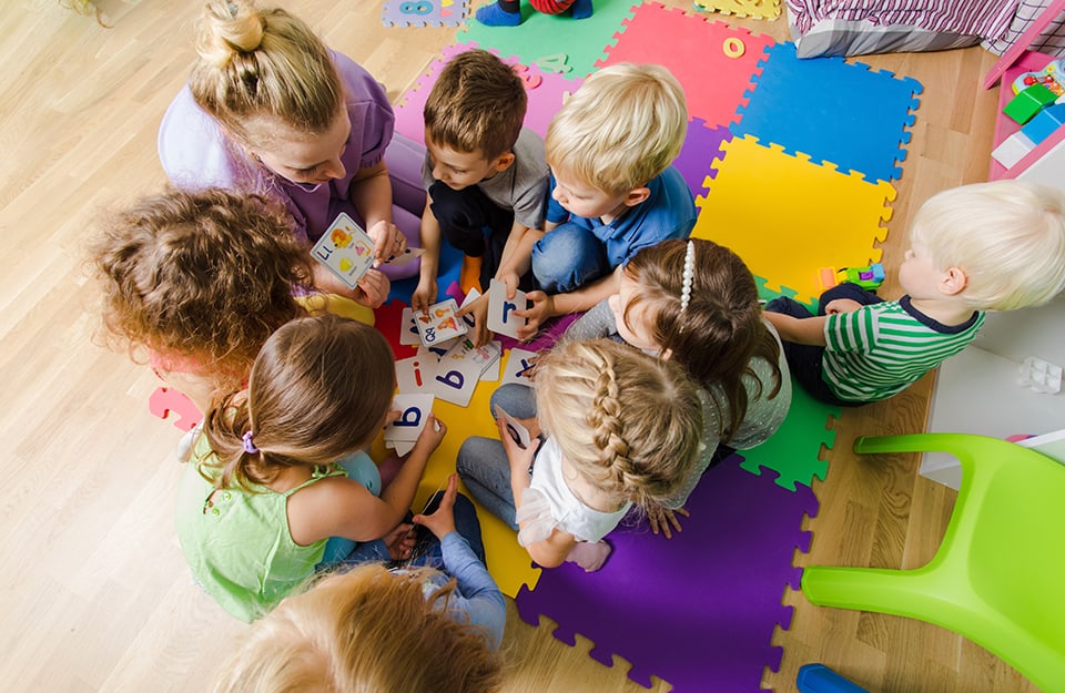 Vista dall'alto di un gruppo di bambine e bambini dell'asilo nido raggruppati in cerchio con una maestra sul parquet della classe, accanto a dei tappetini colorati a forma di puzzle