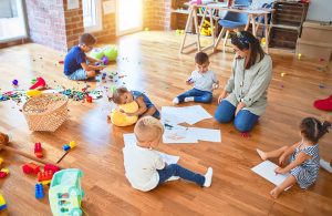 Grande classe di un asilo nido con pavimento in parquet, giocattoli e fogli da disegno sparsi, e una maestra insieme a bambine e bambini che svolgono varie attività