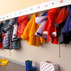 Una fila di appendiabiti in un asilo nido, con cappotti colorati appesi, disegni come segnaposto, stivaletti e una valigetta sul pavimento