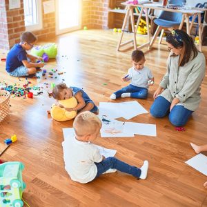Grande classe di un asilo nido con pavimento in parquet, giocattoli e fogli da disegno sparsi, e una maestra insieme a bambine e bambini che svolgono varie attività