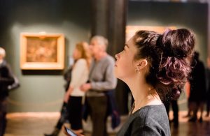 Una ragazza in un museo vista di profilo mentre guarda un quadro fuori campo e dietro di lei si intravedono altre persone