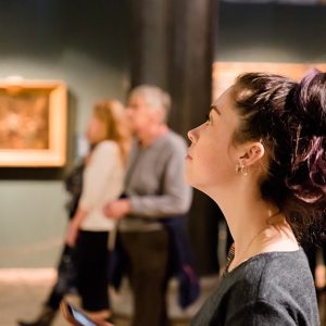 Una ragazza in un museo vista di profilo mentre guarda un quadro fuori campo e dietro di lei si intravedono altre persone