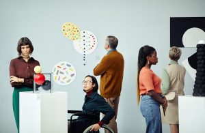 Un gruppo di persone, tra cui una ragazza in sedia a rotella osservano delle sculture e dei quadri di arte astratta nella sala di un museo con pareti e piedistalli bianchi