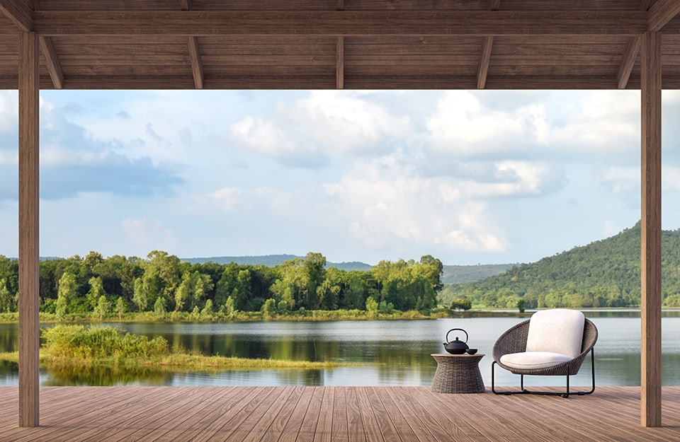 Panorama vista lago da una grande veranda in legno con pavimentazione in legno, una poltrona e un tavolino da caffè in rattan vicino all'acqua. Sul tavolino c'è una teiera