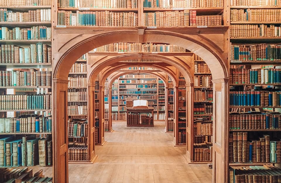 Vista centrale sui numerosi ambienti di una biblioteca pubblica con scaffali in legno disposti attorno a un passaggio ad arco. Al centro, in fondo, si vede un libro antico aperto su un leggio