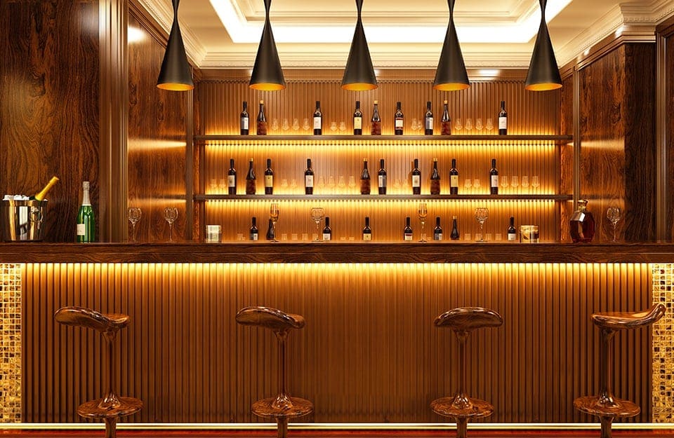 Una sala bar con mobili interamente in legno e soffitto bianco decorato, da cui scendono lampadari conici di design, che illuminano gli sgabelli davanti al bancone, da cui si vedono gli scaffali, pure in legno e illuminati da led, con bottiglie e bicchieri