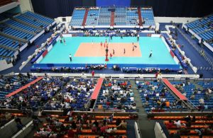 Vista dall'alto e da lontano di un campo di pallavolo sul quale si sta svolgendo una partita, con alcune persone sulle tribune