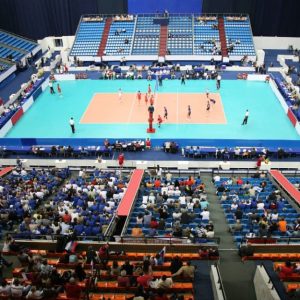 Vista dall'alto e da lontano di un campo di pallavolo sul quale si sta svolgendo una partita, con alcune persone sulle tribune