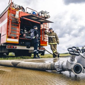 Due Vigili del Fuoco accanto a un'autopompa dalla quale fuoriesce una manichetta con due raccordi