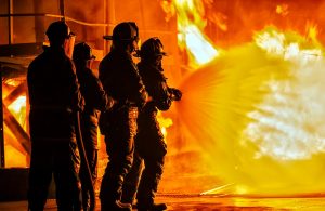 Quattro Vigili del Fuoco altamente equipaggiati sostengono e usano una manichetta provando a domare con l'acqua un grande incendio