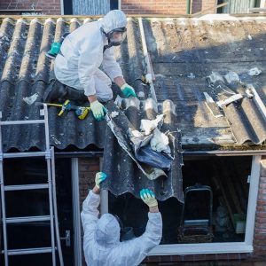 Due operai specializzati nella rimozione di materiali in amianto, protetti da tute speciali, procedono alla rimozione di una lastra ondulata da un tetto