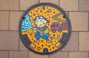 Tipico chiusino giapponese decorato e colorato. Copre un idrante da sottosuolo e vi è rappresentato un pompiere stilizzato che spegne un incendio con la manichetta