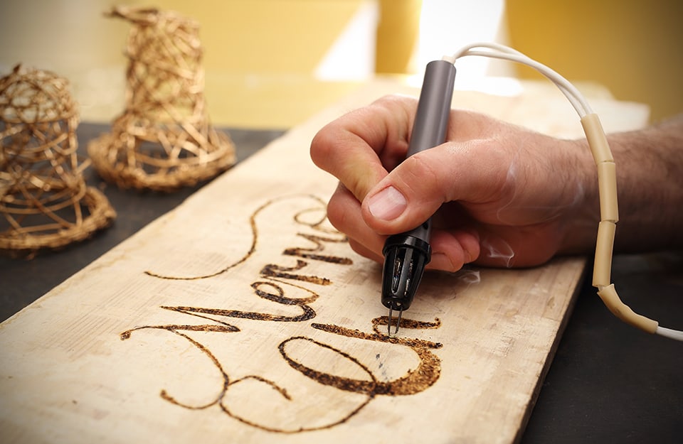 Primo piano sulla mano di un artista che disegna la scritta “Merry Christmas” su legno con una macchinetta elettrica per pirografia