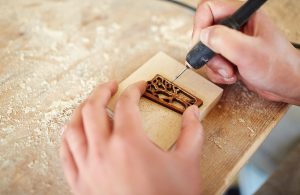 Primo piano sulle mani di un artista che disegna su legno utilizzando una macchina elettrica per pirografia e uno stencil per creare la forma di un albero