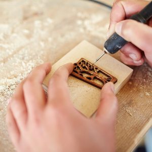 Primo piano sulle mani di un artista che disegna su legno utilizzando una macchina elettrica per pirografia e uno stencil per creare la forma di un albero