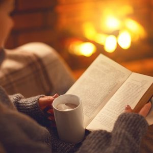 Donna legge un libro in poltrona, davanti al camino, con una tazza di una bevanda calda in mano