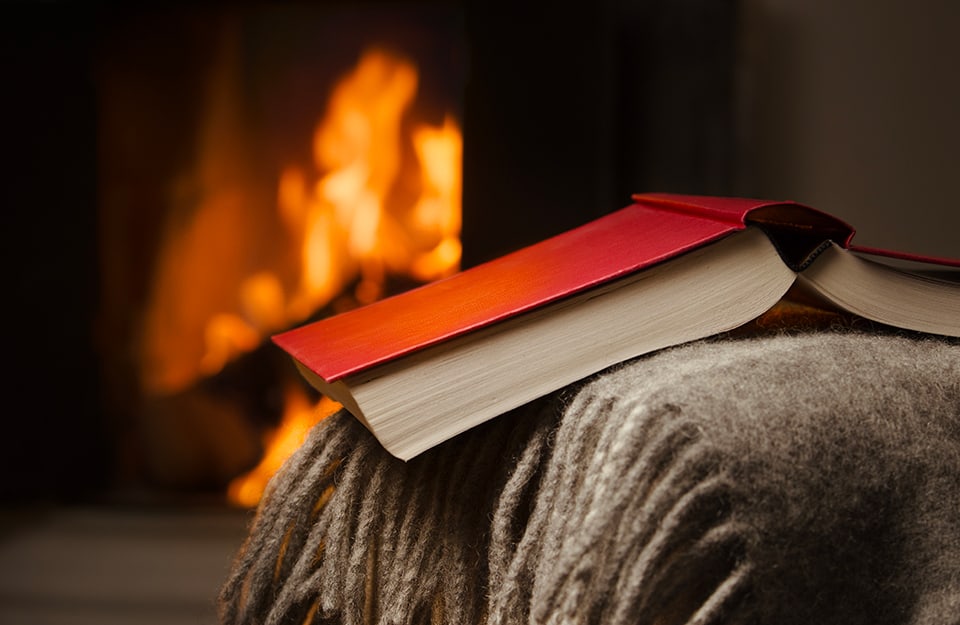 Primo piano di un libro aperto, poggiato sul bracciolo di un divano o di una poltrona, con sotto una coperta. Sullo sfondo un camino con il fuoco acceso