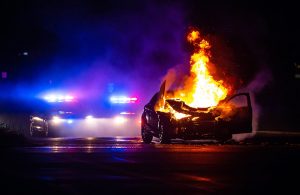Un'auto in fiamme su strada di notte, con sullo sfondo le sirene dei mezzi di soccorso