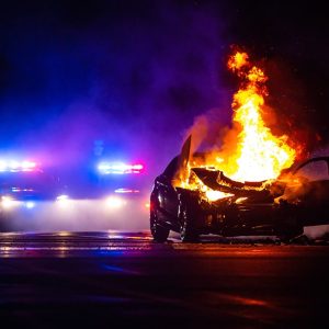 Un'auto in fiamme su strada di notte, con sullo sfondo le sirene dei mezzi di soccorso