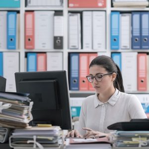 Una donna lavora al computer circondata di faldoni con documenti e alle spalle ha un archivio pieno di raccoglitori