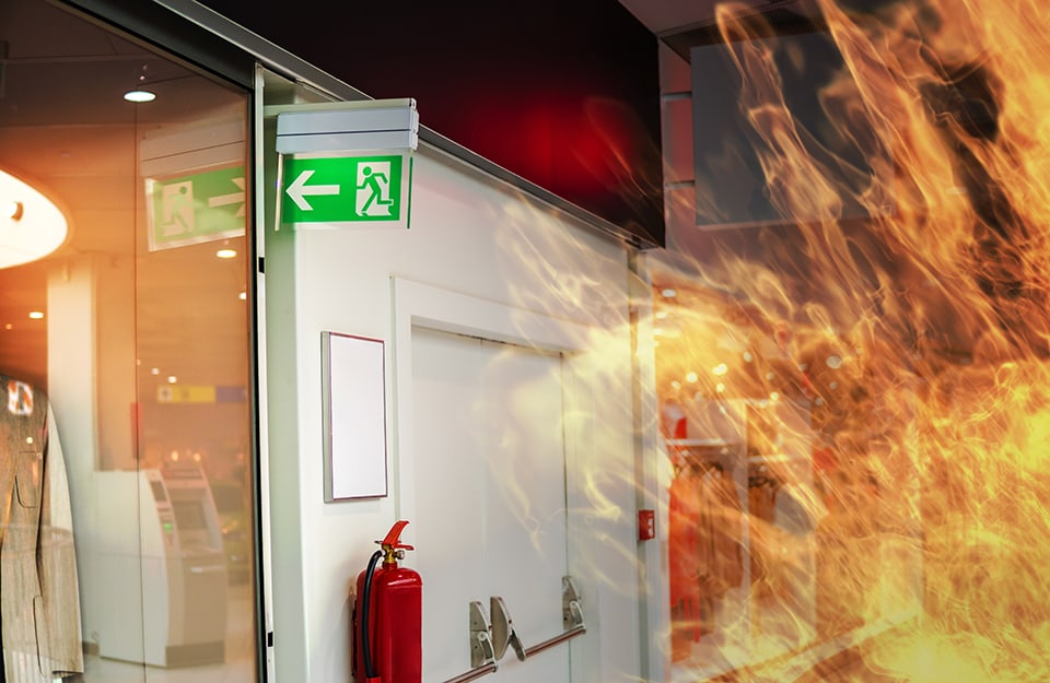 Incendio all'interno di un ufficio in cui si vedono il cartello dell'uscita di sicurezza, un estintore e una porta tagliafuoco con maniglioni