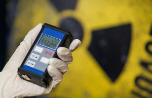 Una mano protetta da un guanto regge un contatore Geiger per misurare il livello di radiazioni, mentre sullo sfondo si vede il simbolo del pericolo radiottivo