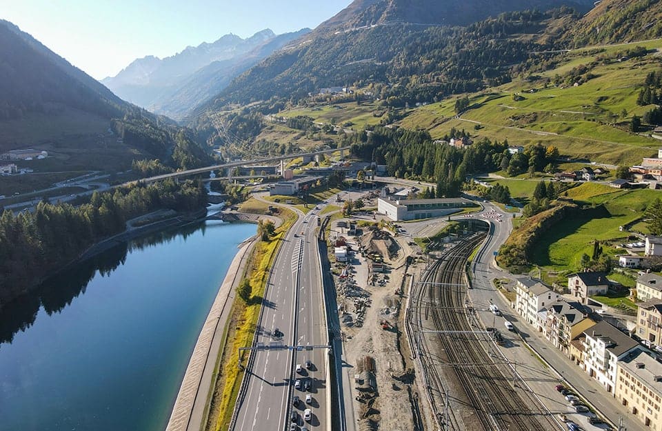 Visuale panoramica dall'alto dell'ingresso al tunnel autostradale e ferroviario del San Gottardo, immerso tra le montagne e parallelo al fiume