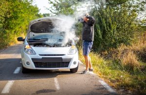 Un uomo sul bordo della strada, in piedi e con le mani in testa, guarda preoccupato la sua auto ferma dalla quale esce fumo dal motore che si vede sotto il cofano alzato