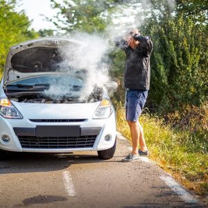 Un uomo sul bordo della strada, in piedi e con le mani in testa, guarda preoccupato la sua auto ferma dalla quale esce fumo dal motore che si vede sotto il cofano alzato