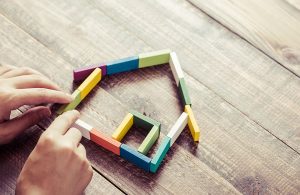 Due mani stanno costruendo con dei mattoncini in legno colorati la forma stilizzata di una casa