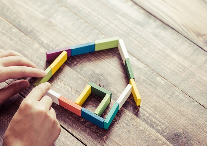 Due mani stanno costruendo con dei mattoncini in legno colorati la forma stilizzata di una casa