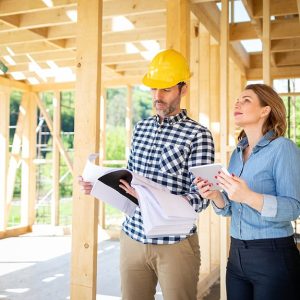 Una donna visita il cantiere di una casa in legno e il capocantiere con casco giallo le insieme a un capo cantiere con una pi