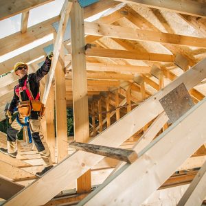 Un operaio edile arrampicato tra le assi di una struttura di un tetto in legno indossando l'equipaggiamento di sicurezza