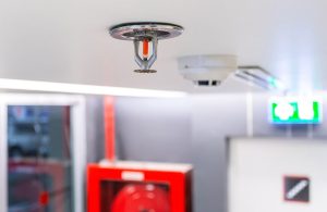 Un sprinkler e un rilevatore di fumi sul soffitto di un locale mentre sullo sfondo di vedono altri dispositivi antincendio, tra cui la segnaletica di sicurezza