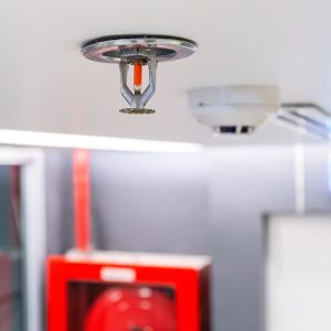 Un sprinkler e un rilevatore di fumi sul soffitto di un locale mentre sullo sfondo di vedono altri dispositivi antincendio, tra cui la segnaletica di sicurezza