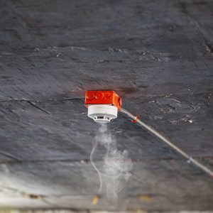 Un rilevatore di fumo bianco e rosso installato su un soffitto in cemento grezzo sta captando del fumo