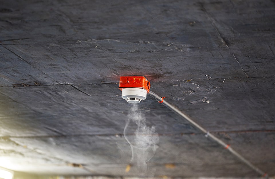 Un rilevatore di fumo bianco e rosso installato su un soffitto in cemento grezzo sta captando del fumo