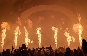 Il palco di un festival musicale, di notte, con il pubblico con le mani alzate e colonne di fuoco che si alzano