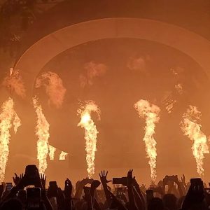 Il palco di un festival musicale, di notte, con il pubblico con le mani alzate e colonne di fuoco che si alzano