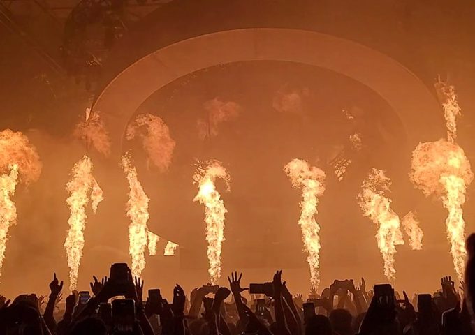 Il palco di un festival musicale, di notte, con il pubblico con le mani alzate e colonne di fuoco che si alzano