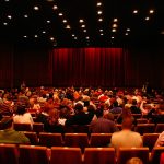 L'affollata platea di un teatro con poltroncine rosse e il sipario chiuso, anche questo rosso