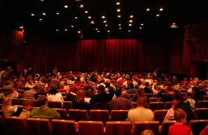 L'affollata platea di un teatro con poltroncine rosse e il sipario chiuso, anche questo rosso