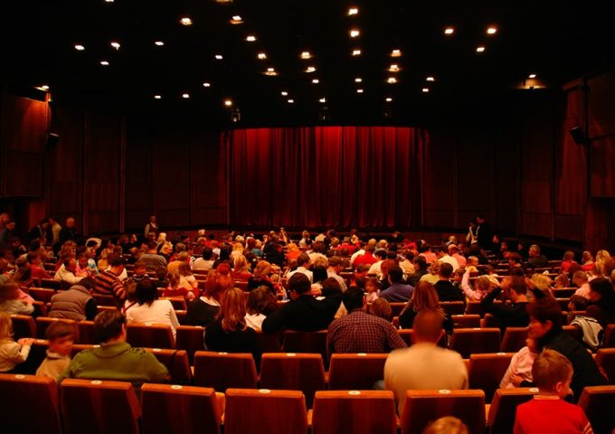 L'affollata platea di un teatro con poltroncine rosse e il sipario chiuso, anche questo rosso