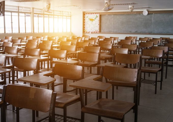 Vista di tre quarti di un'aula scolastica senza alunni ma piena di banchi singoli in legno, con la luce del sole, all'alba o al tramonto, che entra dalla lunga finestra