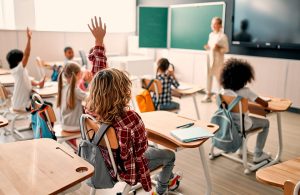 Una classe piena di bambine e bambini, alcuni dei quali stanno alzando la mano, mentre sullo sfondo si vede la maestra davanti a delle lavagne e a uno schermo