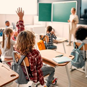 Una classe piena di bambine e bambini, alcuni dei quali stanno alzando la mano, mentre sullo sfondo si vede la maestra davanti a delle lavagne e a uno schermo