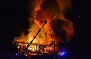 Vista da lontano di alcuni mezzi pesanti dei vigili del fuoco che tentano di domare un incendio in un grande edificio ridotto ormai a uno scheletro