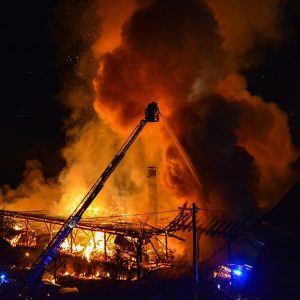 Vista da lontano di alcuni mezzi pesanti dei vigili del fuoco che tentano di domare un incendio in un grande edificio ridotto ormai a uno scheletro